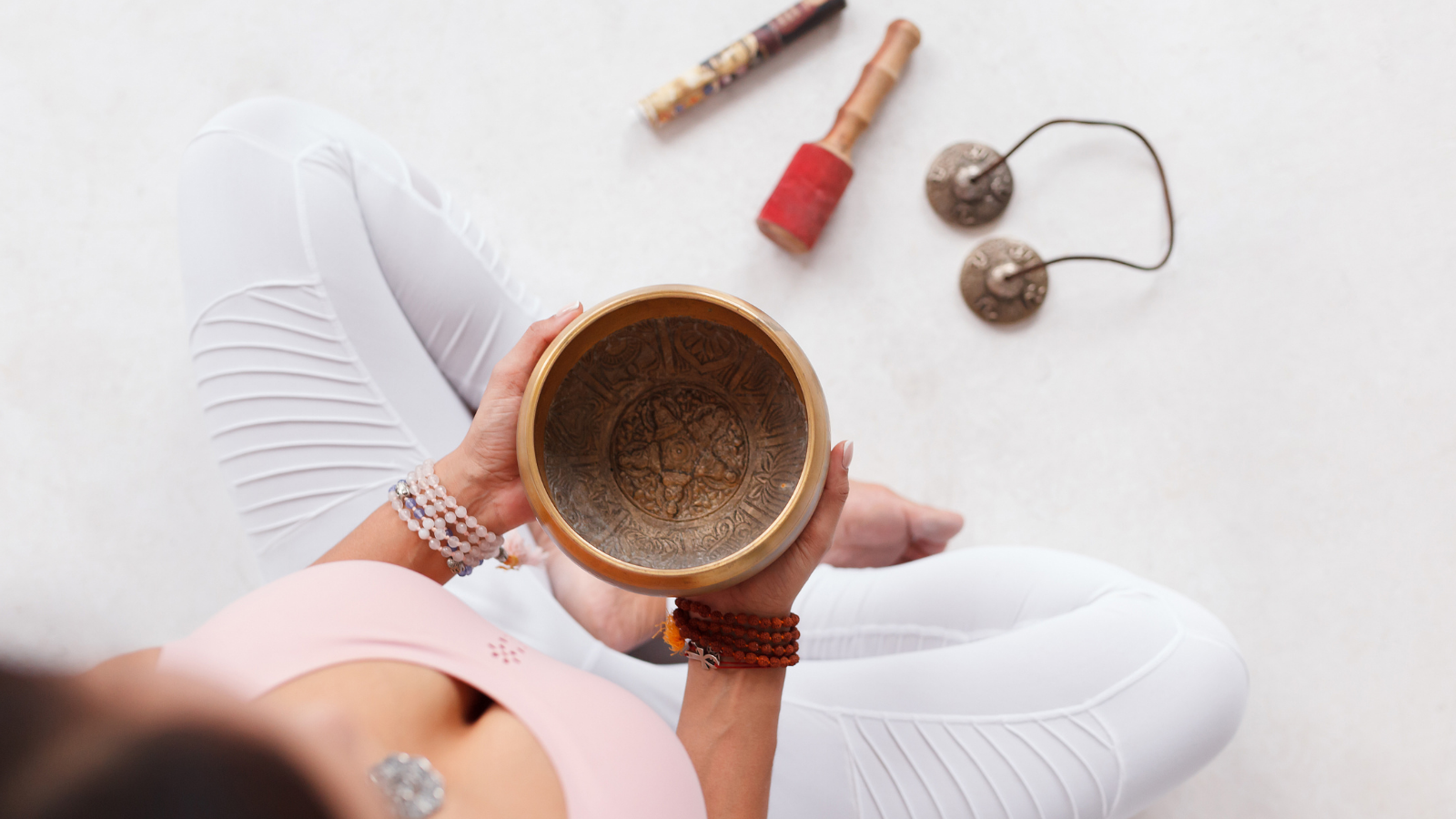 Unknown young woman holding Tibetan singing bowl and pestle sitting on the floor next to mortar and fragrant candles for yoga and meditation.