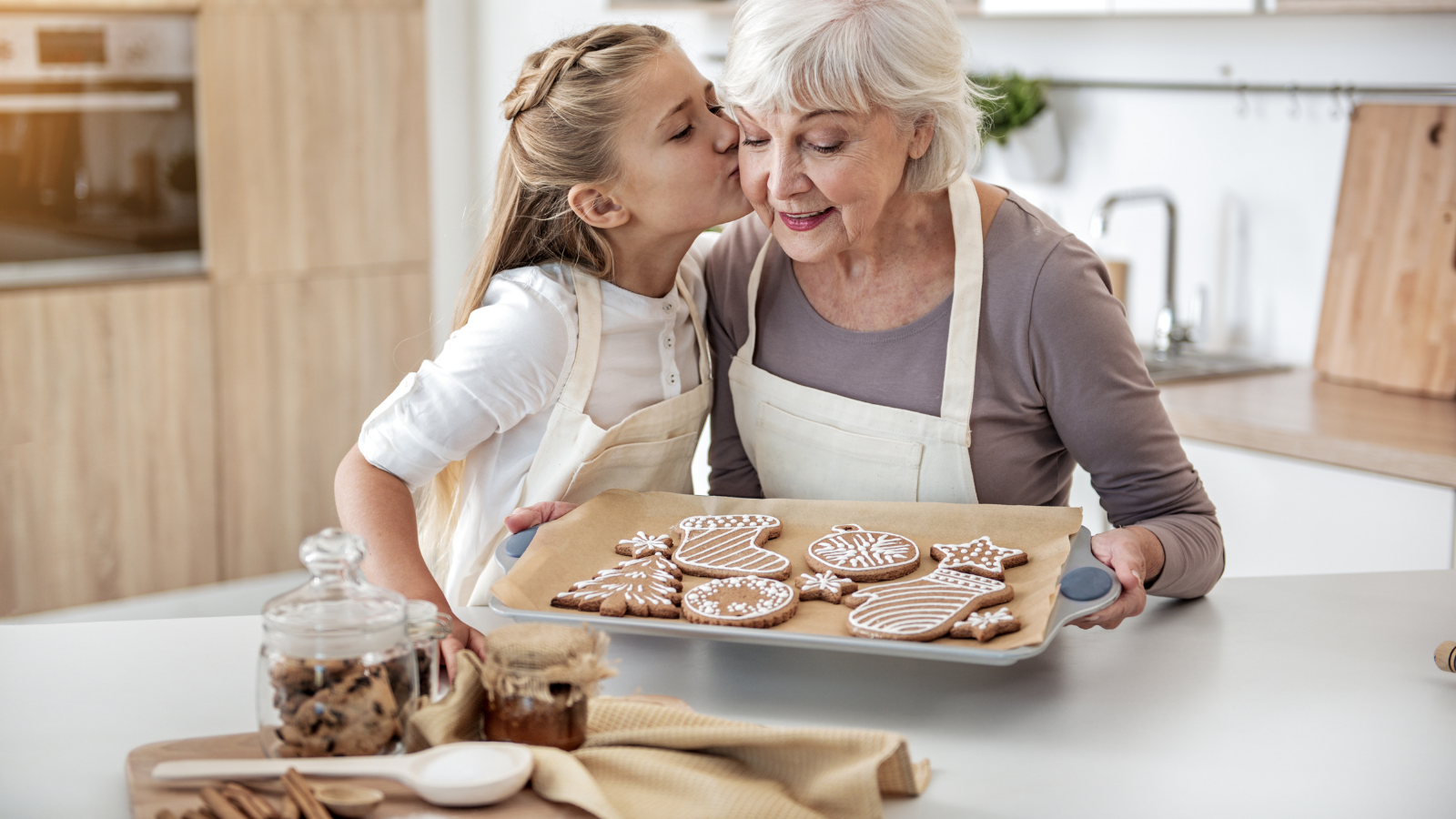 Cute girl is kissing cheek of her grandmother with love. Senior woman is holding pray with self-baked cookies and smiling.