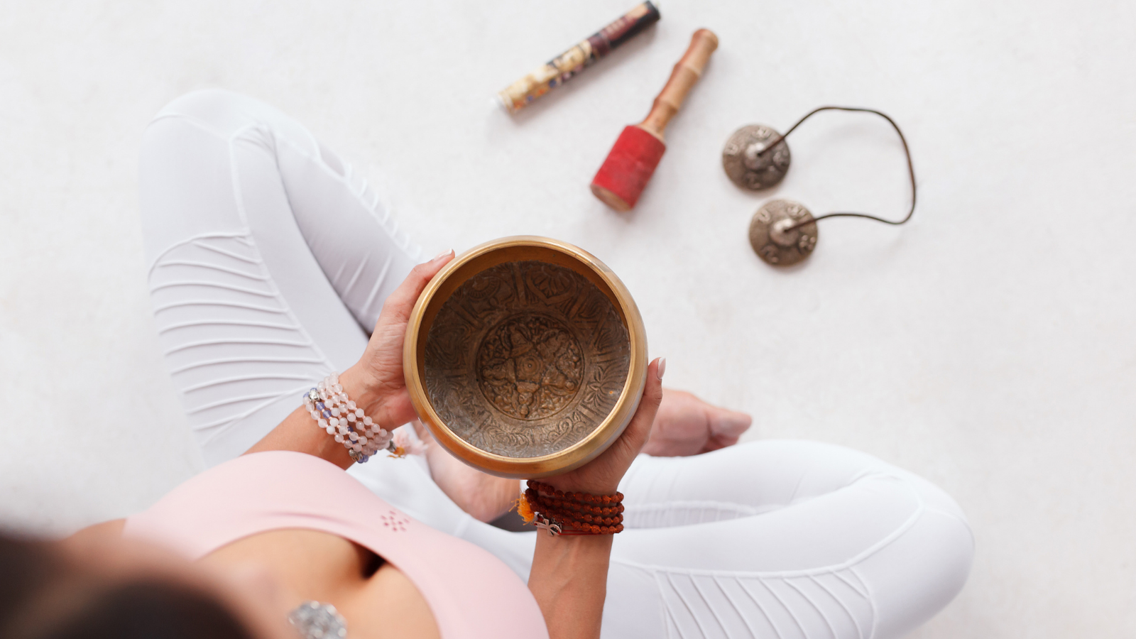 Young woman holding Tibetan singing bowl and pestle sitting on the floor next to mortar and fragrant candles for yoga and meditation