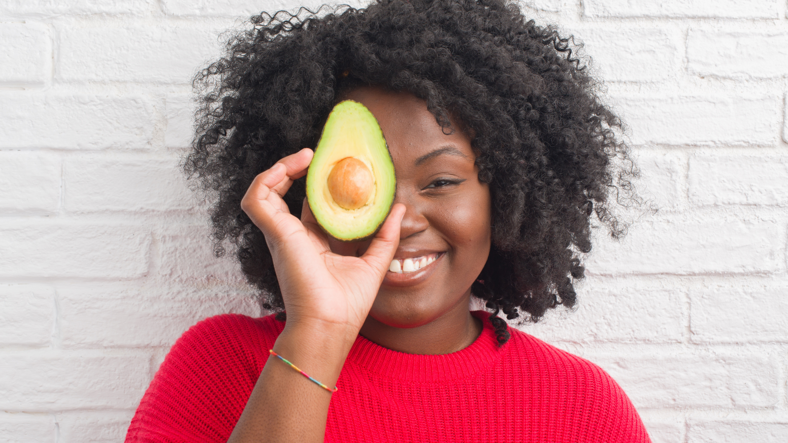 Young woman by white brick wall eating avocado with a happy face standing and smiling with a confident smile