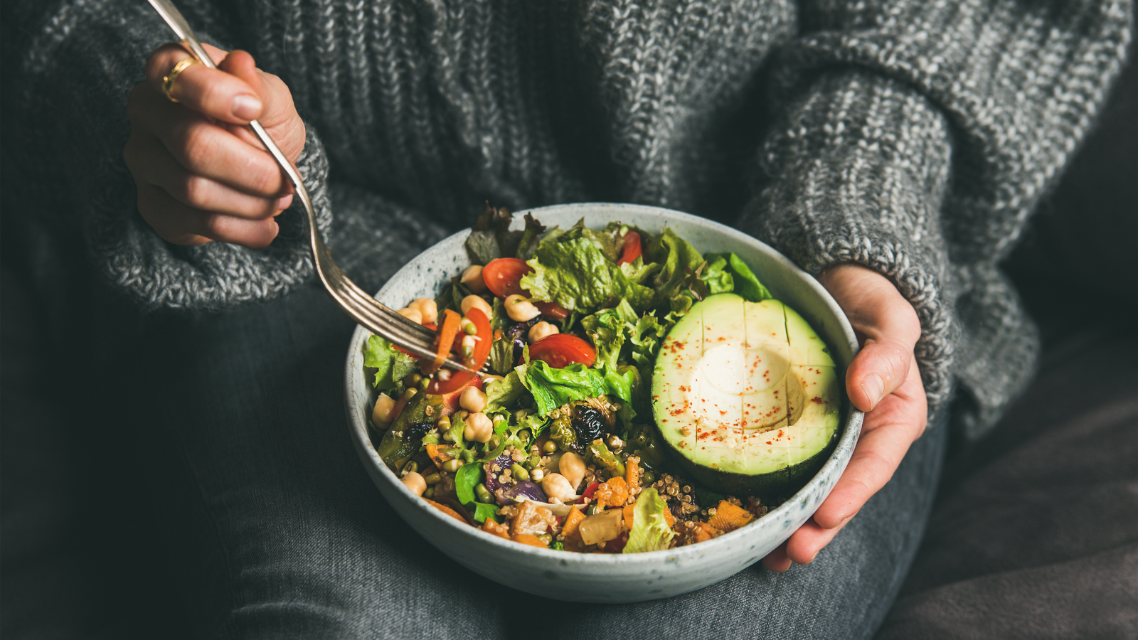 Healthy vegetarian dinner. Woman in jeans and warm sweater holding bowl with fresh salad, avocado, grains, beans, roasted vegetables.