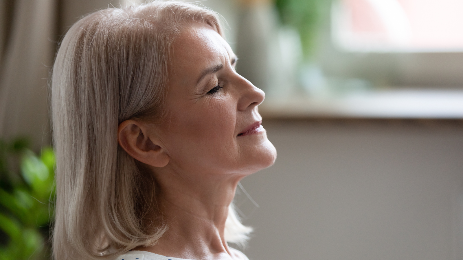 Calm serene middle aged woman meditating with eyes closed inhaling fresh air relaxing indoors