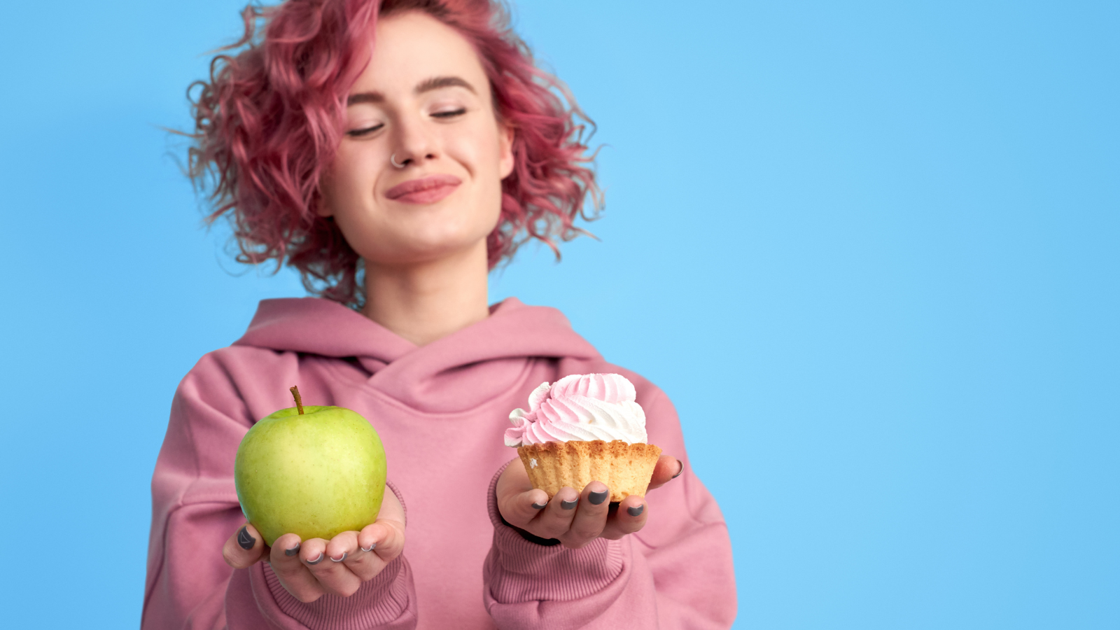 Pretty smiling girl with pink hair in hoodie comparing between fresh green apple and sweet cupcake in hands over blue background