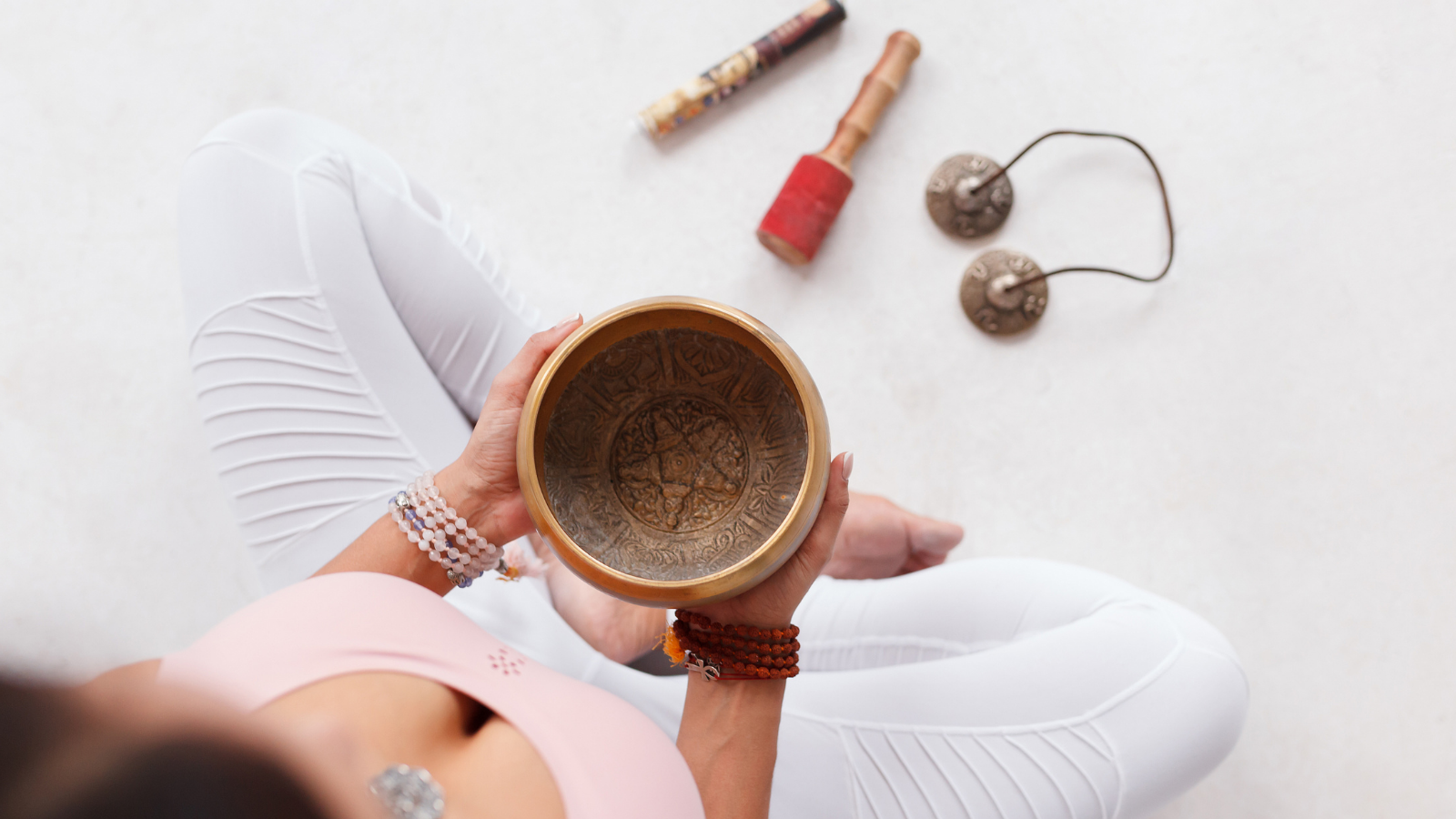 Unknown young woman holding Tibetan singing bowl and pestle sitting on the floor next to mortar and fragrant candles for yoga and meditation