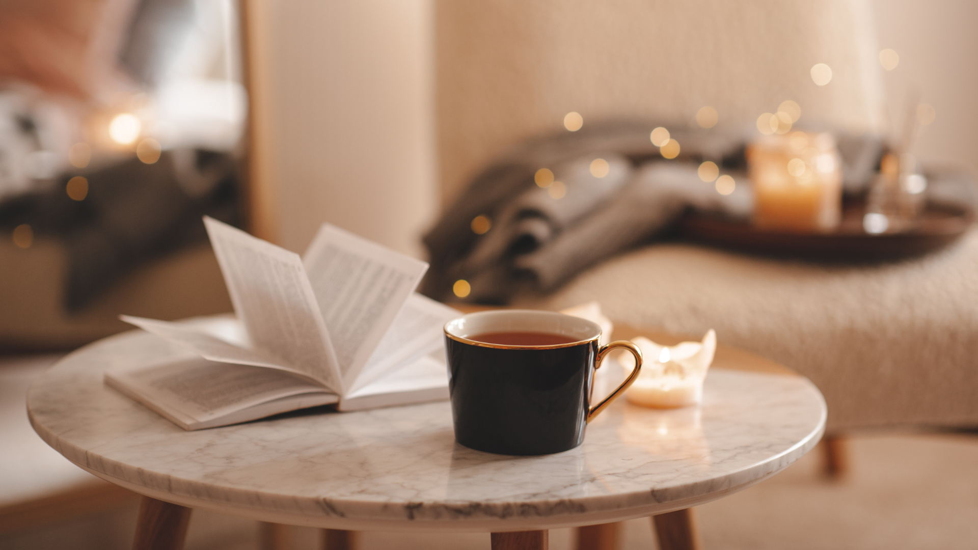 Cup of tea with paper open book and burning scented candles on marble table over cozy chair and glowing lights in bedroom closeup. Winter holiday season.