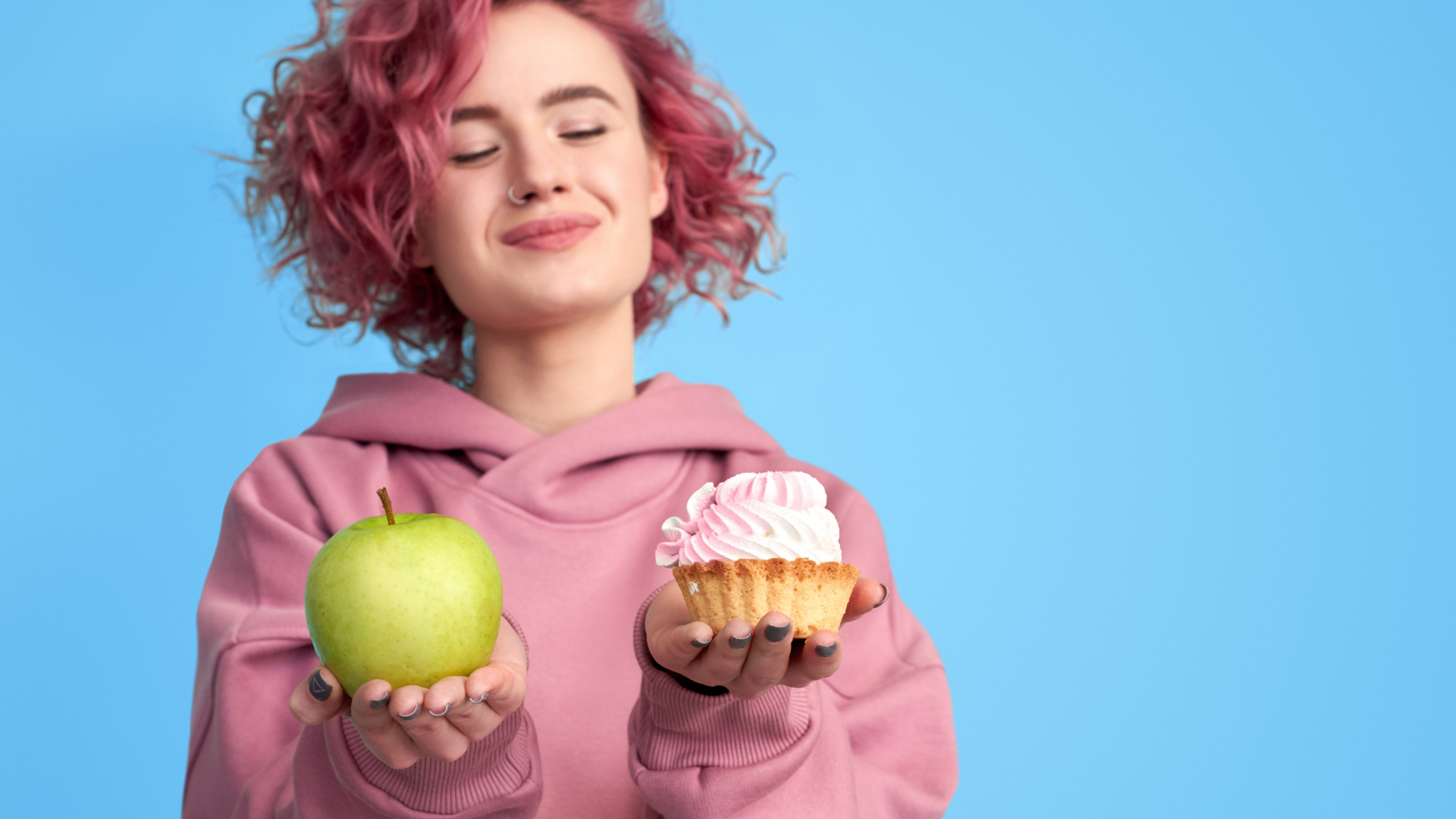 Pretty smiling girl with pink hair in hoodie comparing between fresh green apple and sweet cupcake.