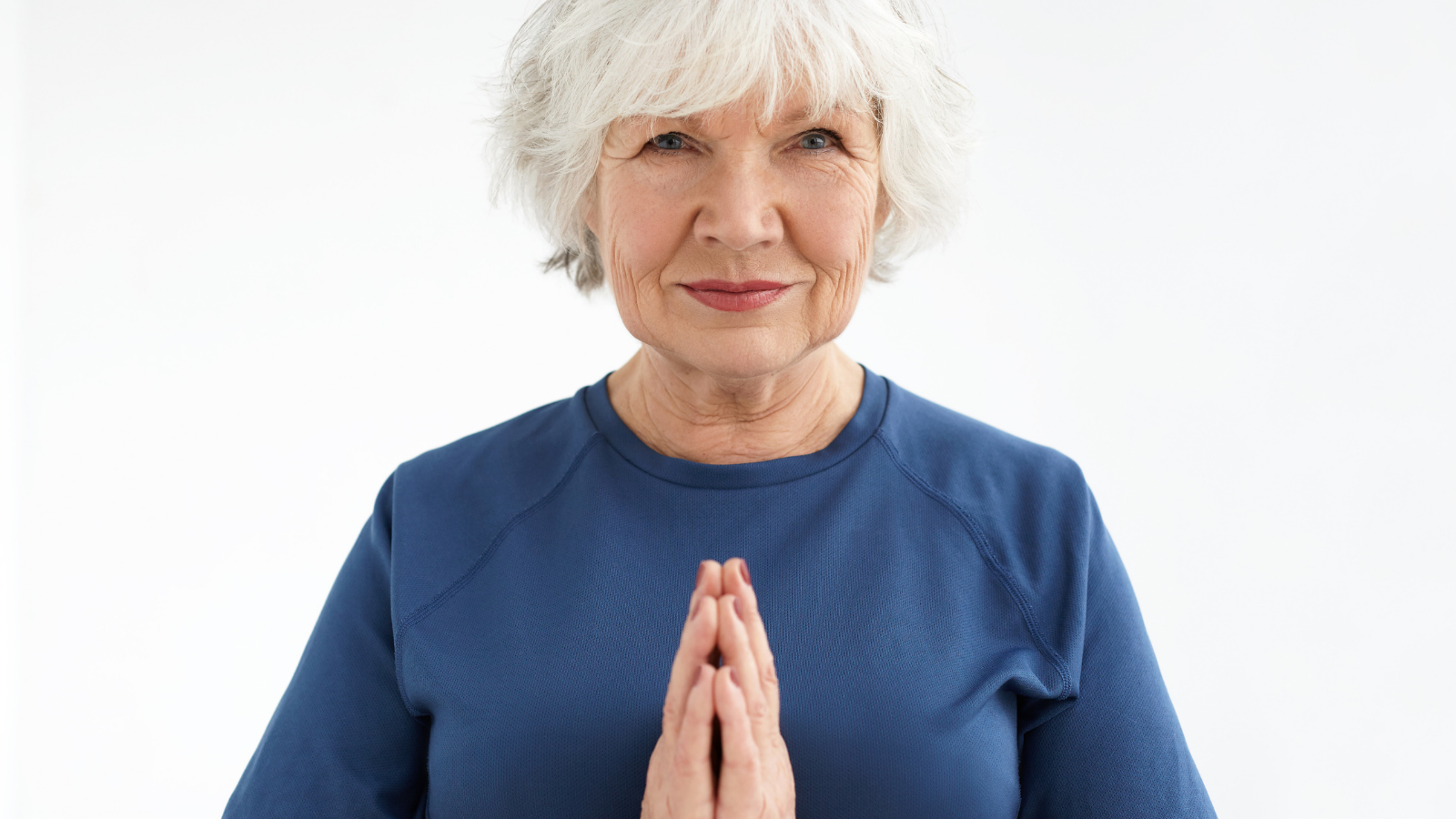 Calm and beautiful elderly woman with short gray hair and wrinkles holding hands in namaste during yoga practice.