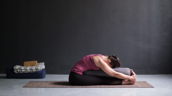 Female yoga student practicing seated forward bend (paschimottanasana) with yoga bolster for support. 