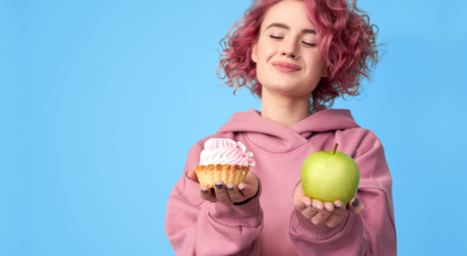 Pretty smiling girl with pink hair in hoodie comparing between fresh green apple and sweet cupcake i