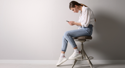 Woman with bad posture using smartphone while sitting on stool near light grey wall.