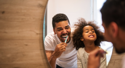 Father and daughter happily brushing teeth together in the mirror.