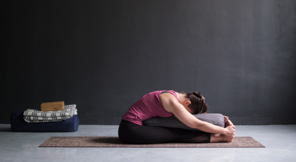 Female yoga student practicing seated forward bend (paschimottanasana) with yoga bolster for support. 