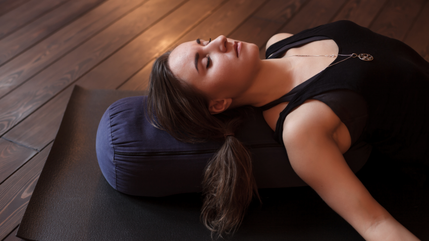 How to practice Corpse Pose or Savasana for greater comfort and ease Woman resting after a yoga class in Corpse Pose or Savasana with bolster for spine comfort