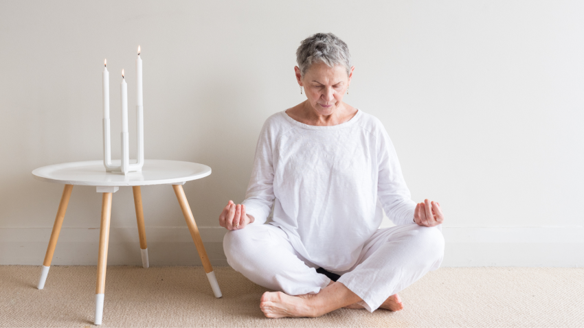 woman in white sitting in yoga meditation position with candles against neutral wall background
