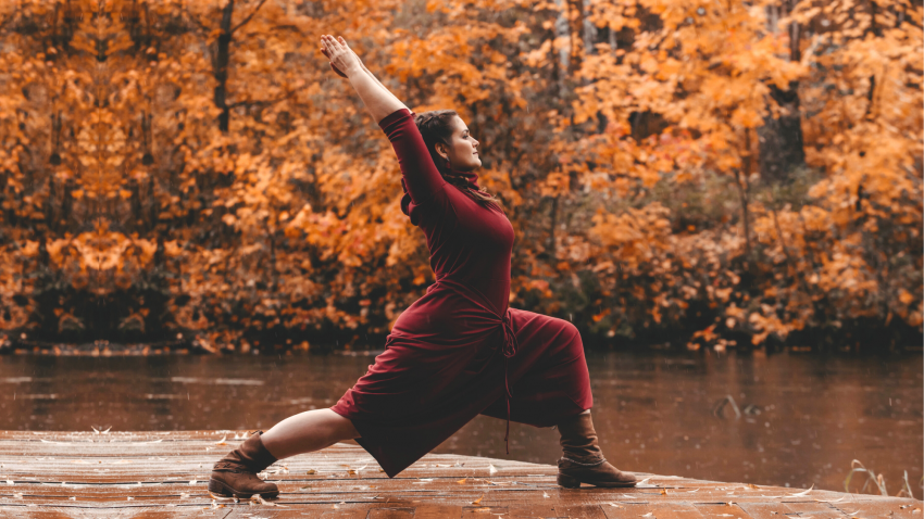 curvy woman practicing warrior 1 on dock near lake and autumn fall leaves